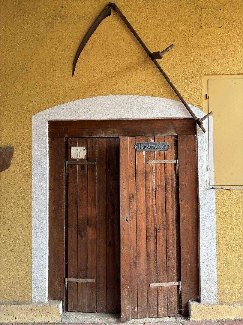 Gelbe Hauswand mit rustikaler Holztür und einer Sense als Dekoration darüber.