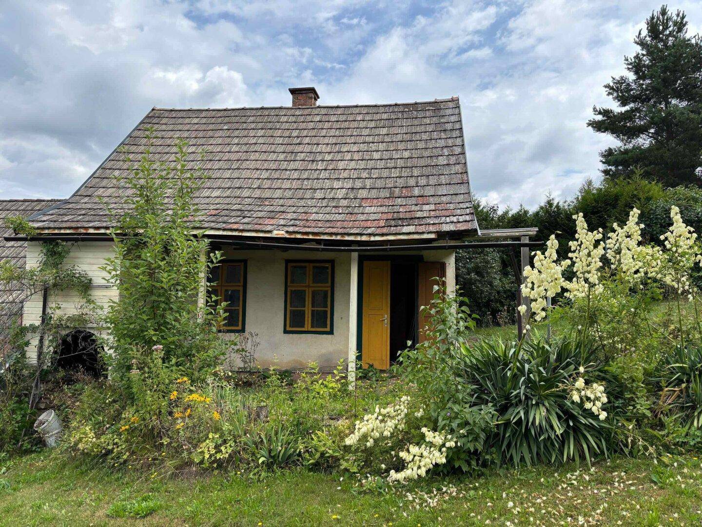 Altes Landhaus umgeben von üppiger Vegetation und hohen Bäumen unter bewölktem Himmel.