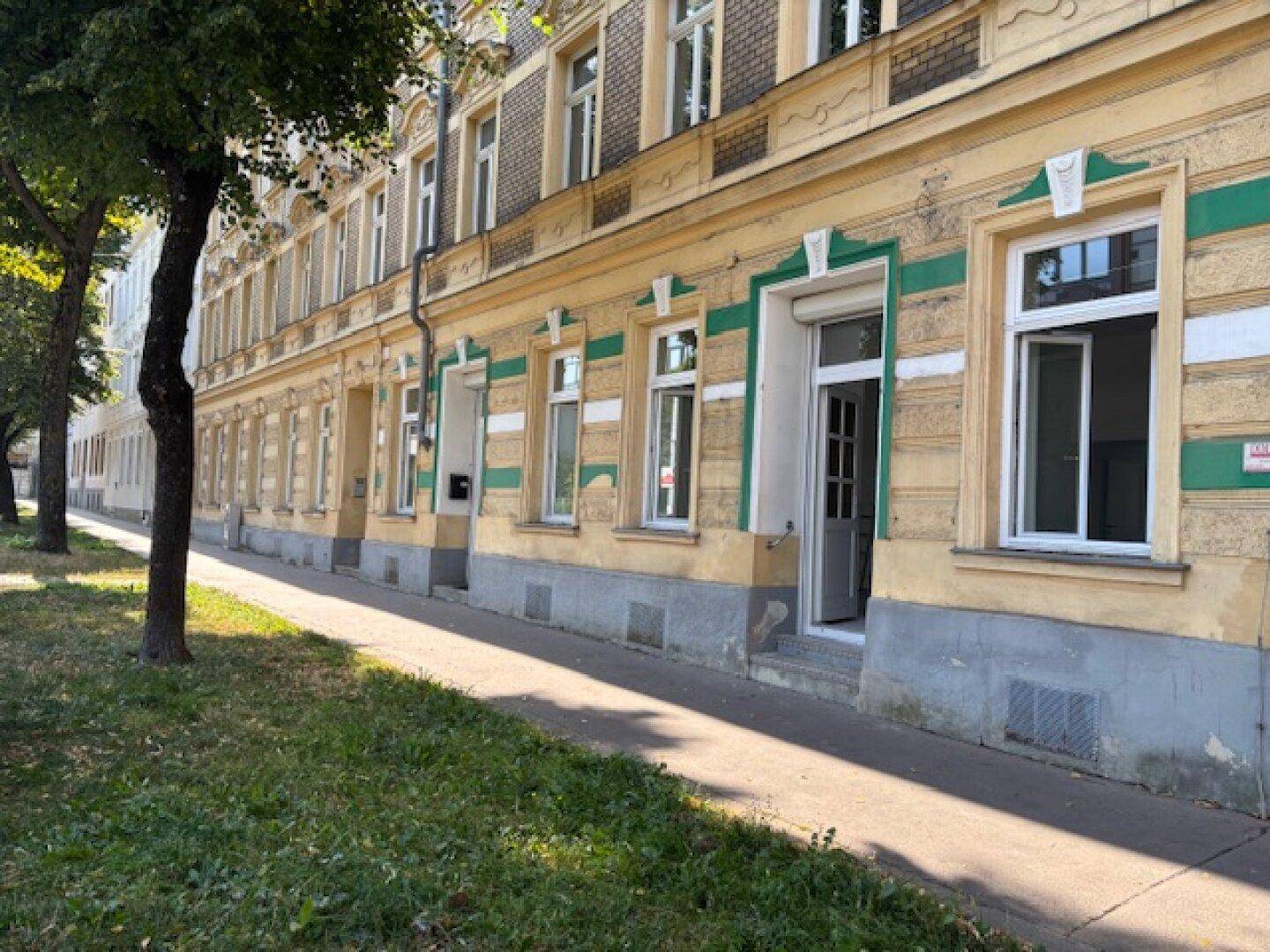 Fassade eines Altbaus mit gelb-grüner Farbgebung und Bäumen am Bürgersteig.