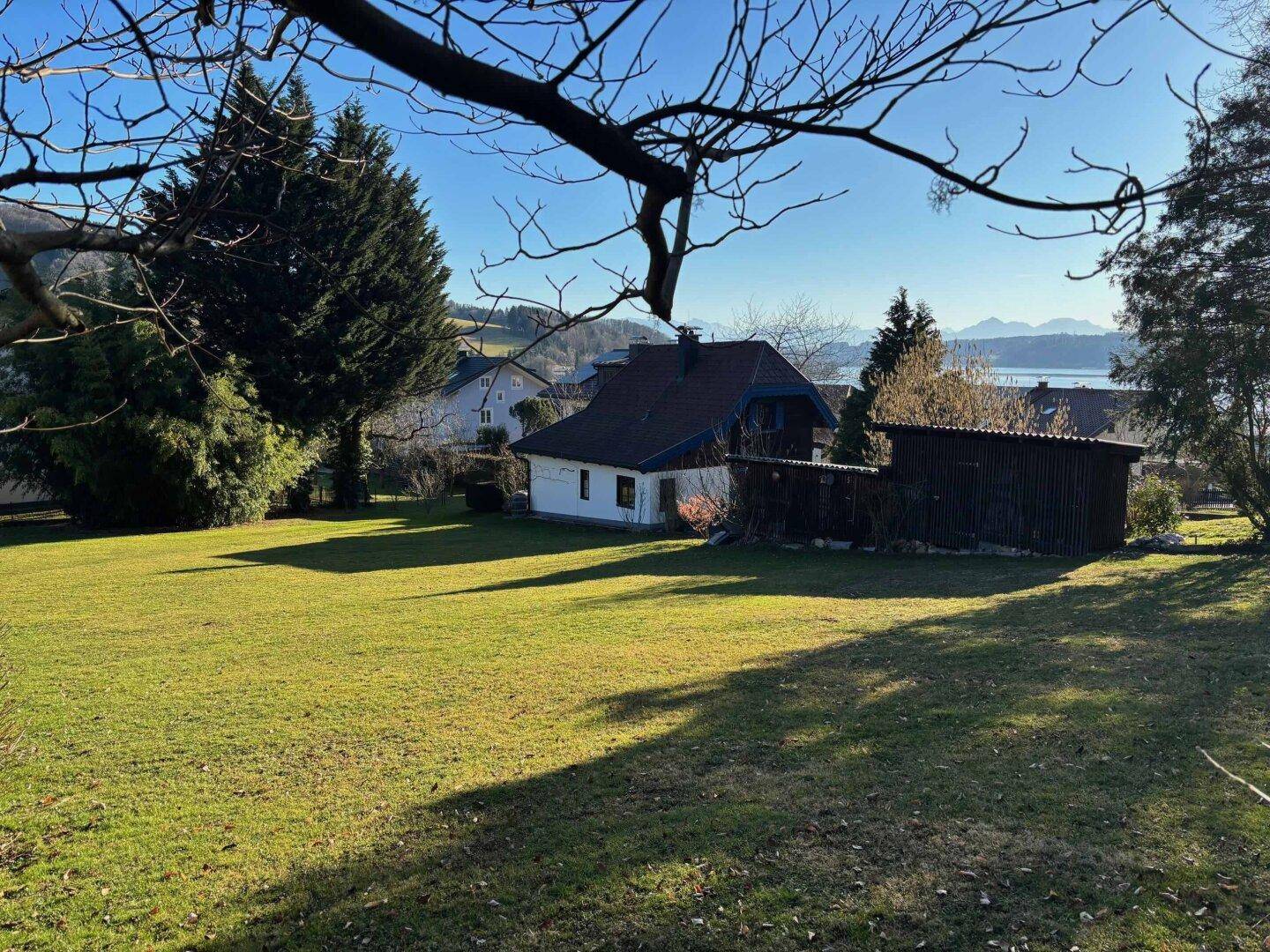 Sonnige Rasenfläche mit einem kleinen Haus im Hintergrund und einem weiten Blick auf den See und die Berge.