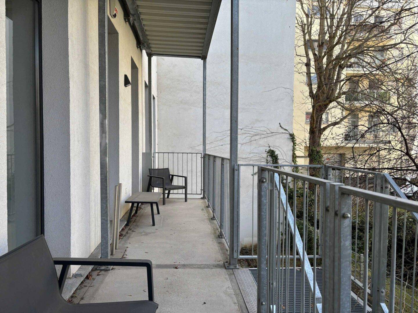 Langer, schmaler Balkon mit Metallgeländer und einfacher Bestuhlung, umgeben von Bäumen.