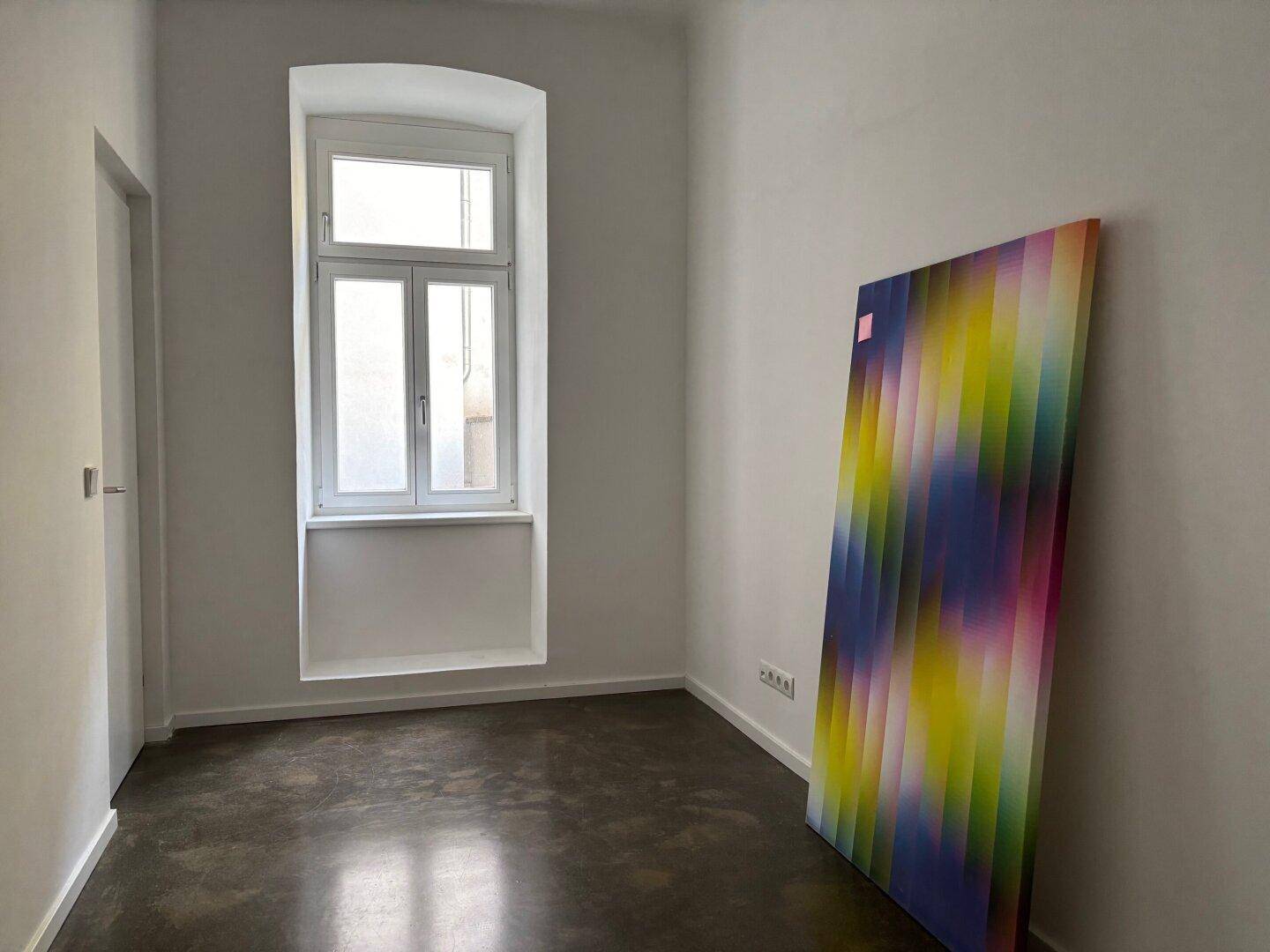 Kleines Zimmer mit einem hohen Fenster, das viel Tageslicht hereinlässt, und einem farbenfrohen Kunstwerk.