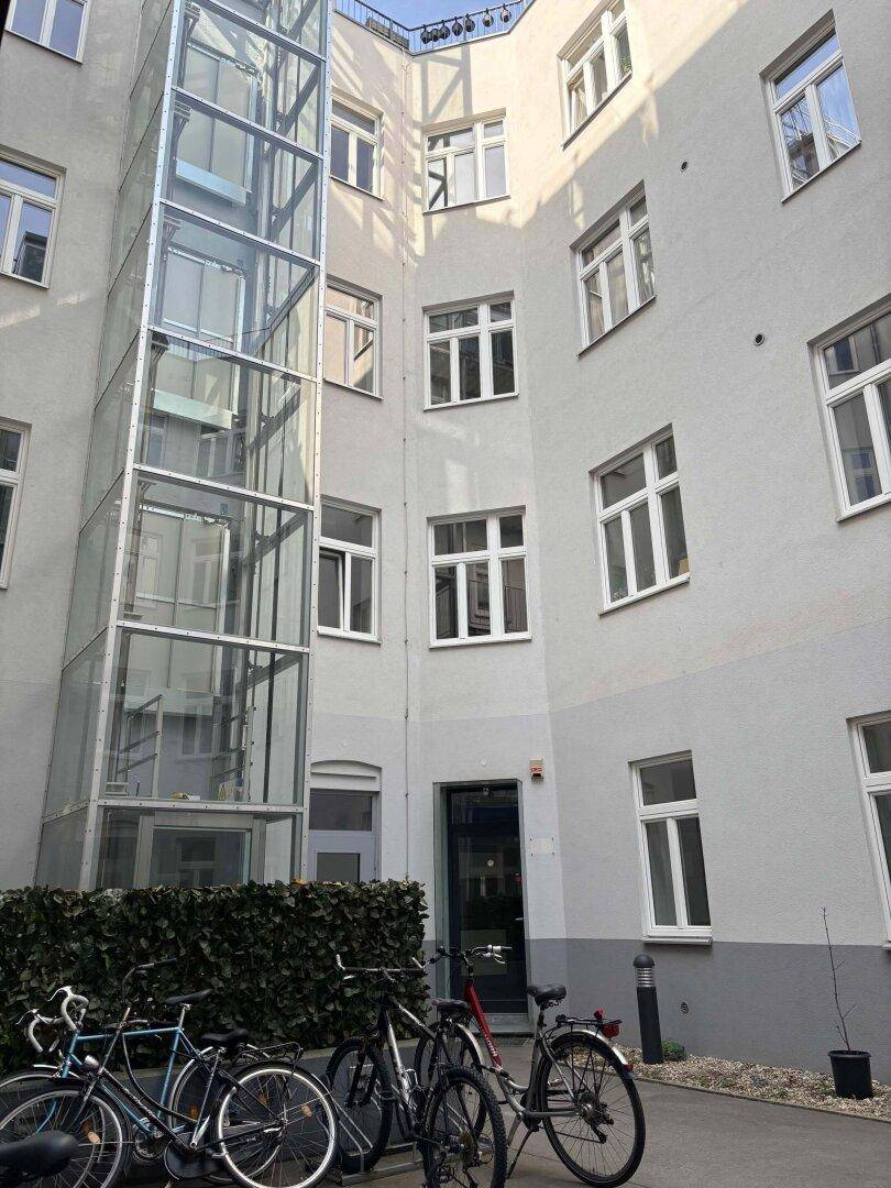 Innenhof eines Gebäudes mit einem modernen Glasaufzug und abgestellten Fahrrädern im Vordergrund.