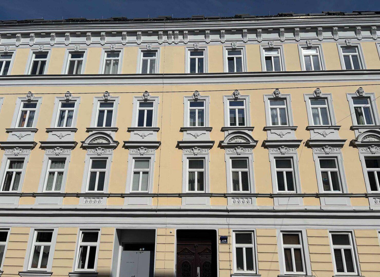 Historische Fassade eines Altbaus mit vielen Fenstern und dekorativen Stuckelementen unter blauem Himmel.