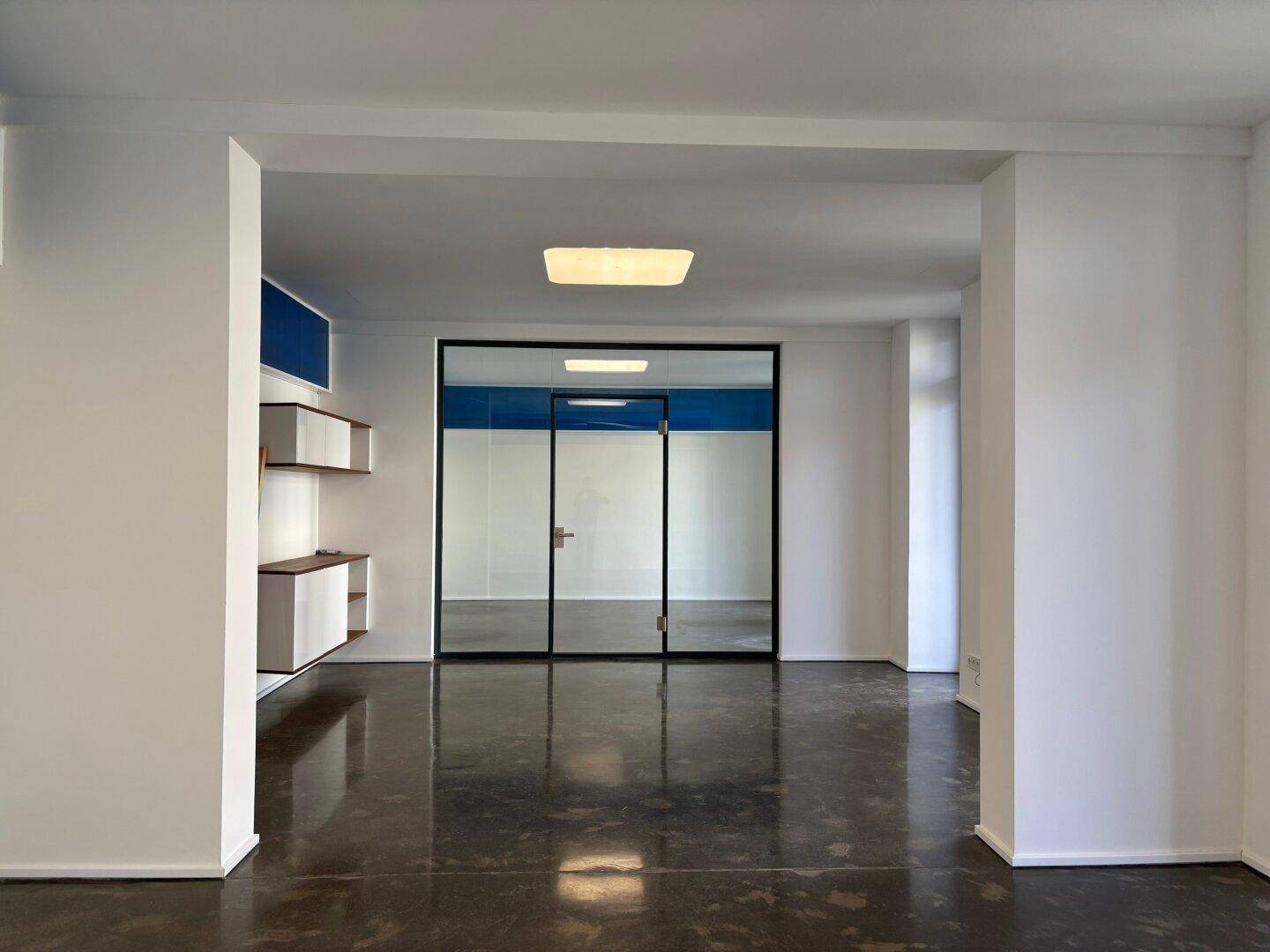 Heller Flur mit dunklem Betonboden und einer modernen Glastür, die zu einem weiteren Raum führt.