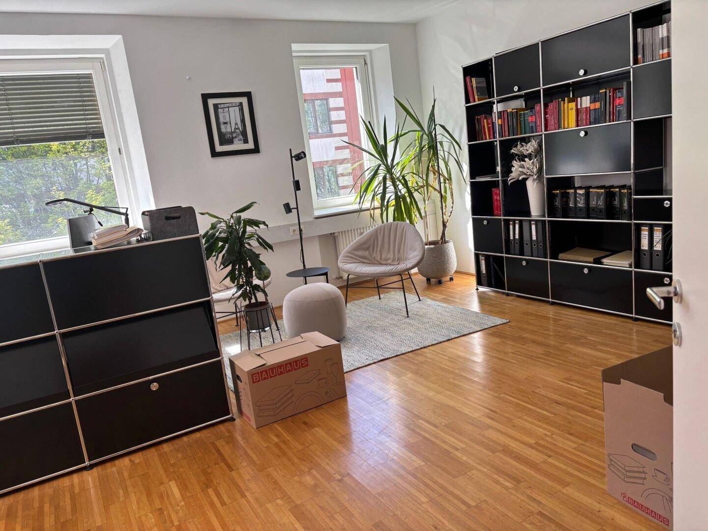 Helles Büro mit Holzparkett, schwarzen modularen Regalsystemen und bequemen Sitzgelegenheiten.