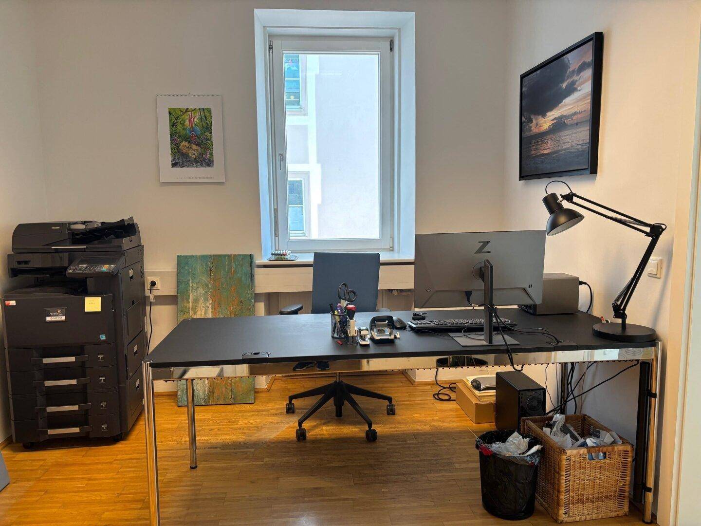 Arbeitsplatz mit großem Schreibtisch, Computer, Drucker und einem Fenster, das viel Licht hereinlässt.