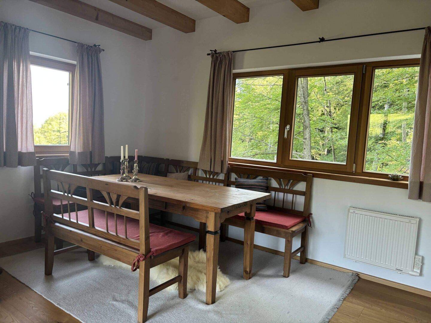 Rustikaler Essbereich mit Holztisch, Bänken und Fenstern mit Blick ins Grüne.