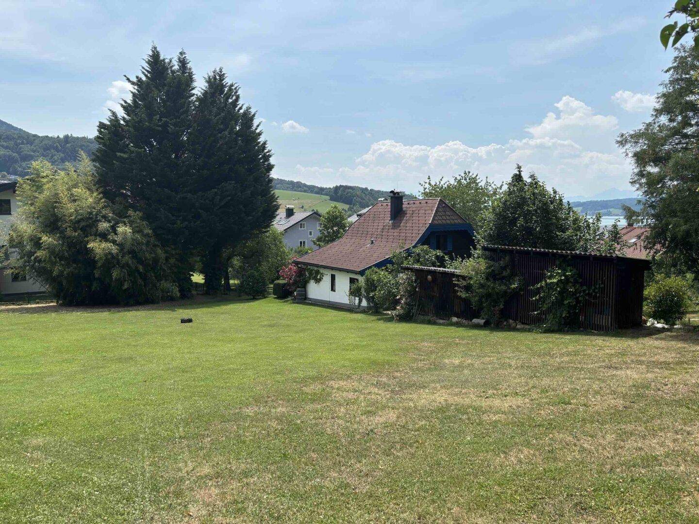 Weitläufiger Rasenbereich mit einem Haus und Schuppen im Hintergrund, umgeben von Bäumen und mit Seeblick.