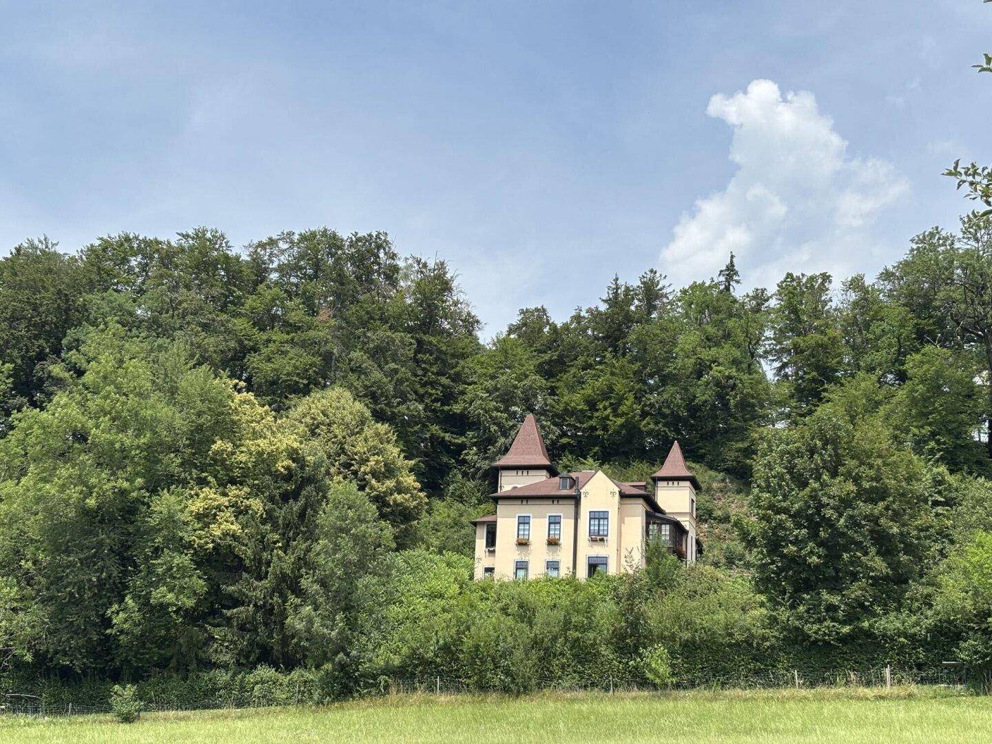 Ein markantes gelbes Haus mit Türmchen, eingebettet in eine bewaldete Hügellandschaft unter blauem Himmel.