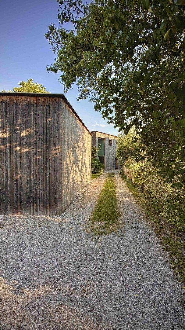 Langer Kiesweg führt zu einem modernen Holzhaus, umgeben von Bäumen und Sträuchern.