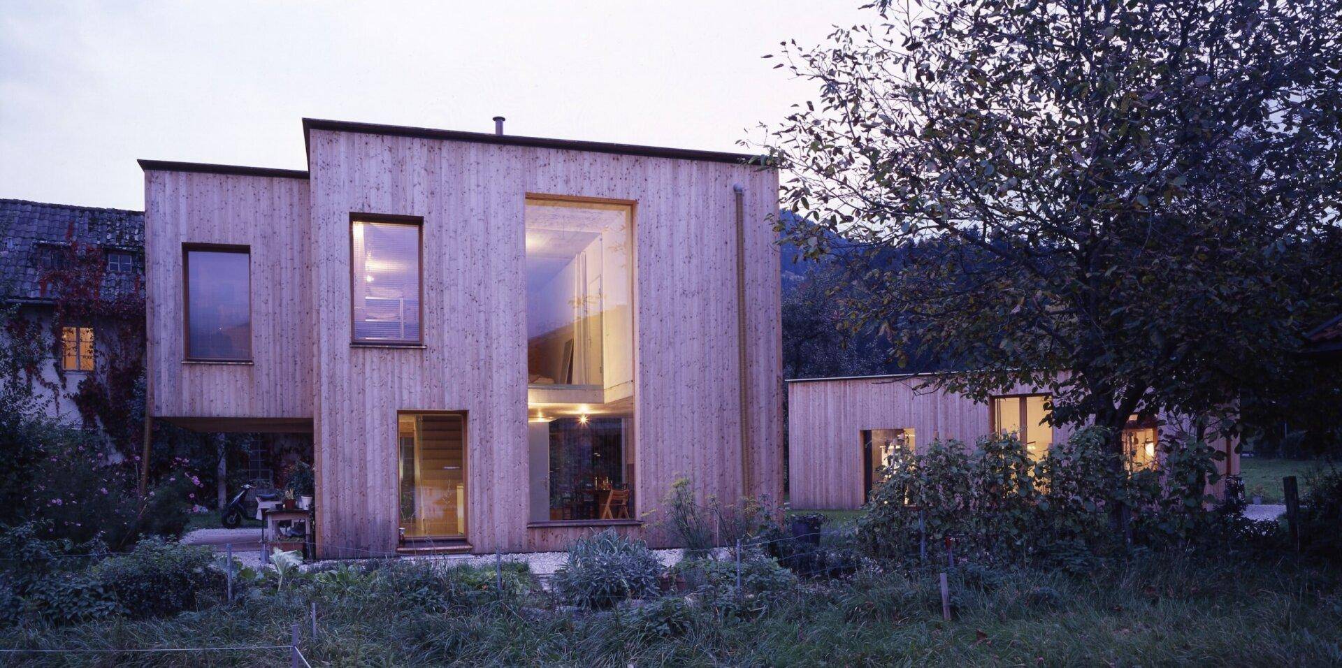 Modernes Holzhaus in der Dämmerung mit beleuchteten Innenräumen und großen Glasflächen.