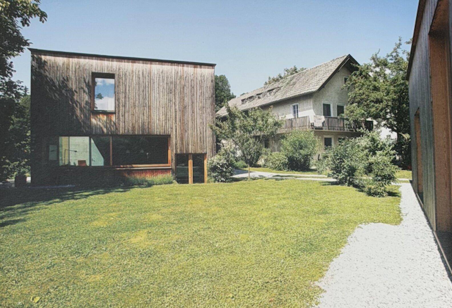 Modernes Holzgebäude mit großem Rasen und einem traditionellen Haus im Hintergrund.