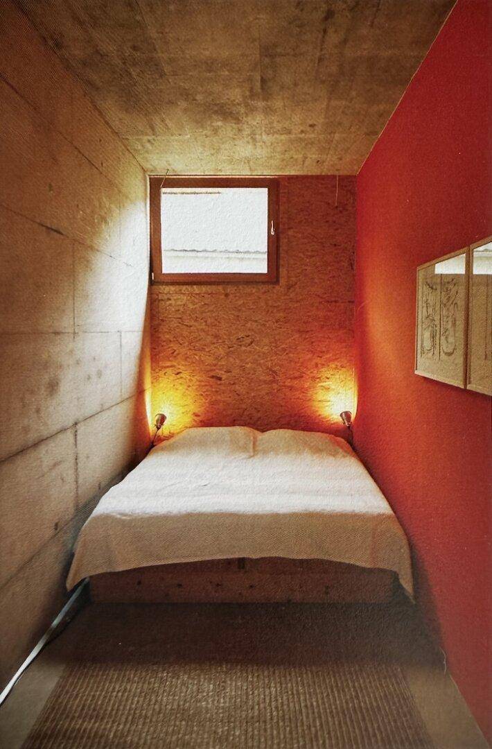 Gemütliches Schlafzimmer mit Holzwänden, einer roten Akzentwand und einem kleinen Fenster.