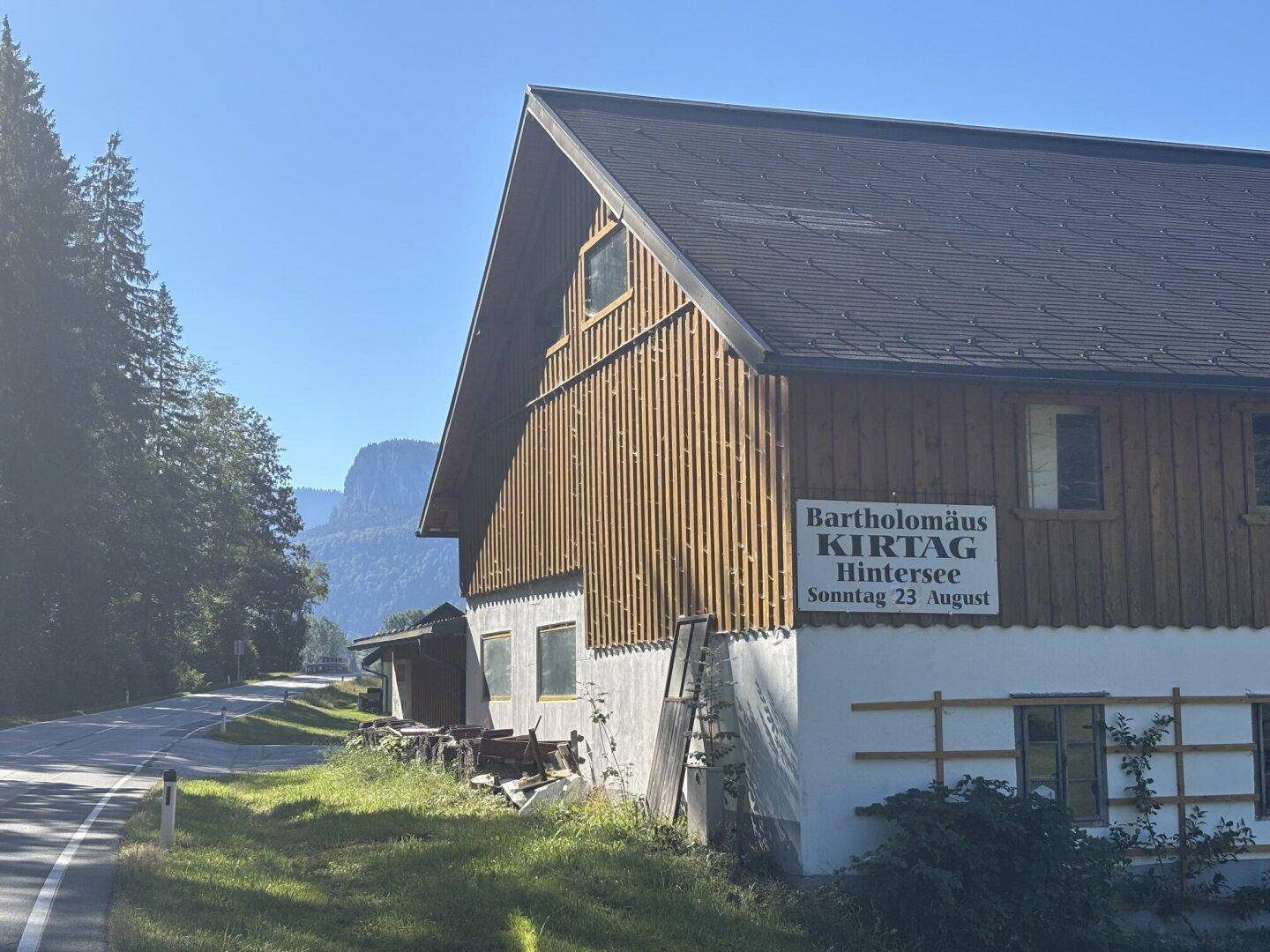 Traditionelles Holzgebäude mit einem Veranstaltungshinweis an einer Landstraße in den Bergen.