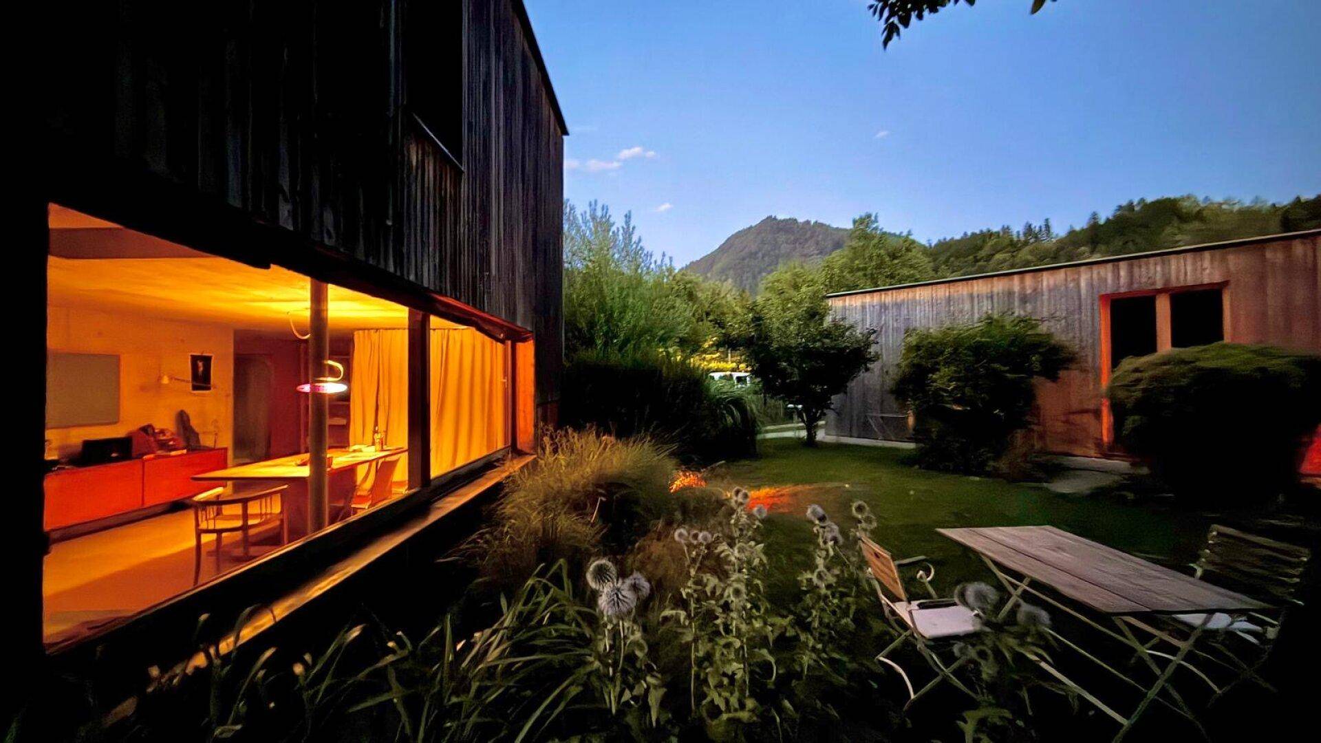 Modernes Holzhaus mit großem Fenster, gemütlichem Garten-Sitzbereich und Bergblick in der Abenddämmerung.