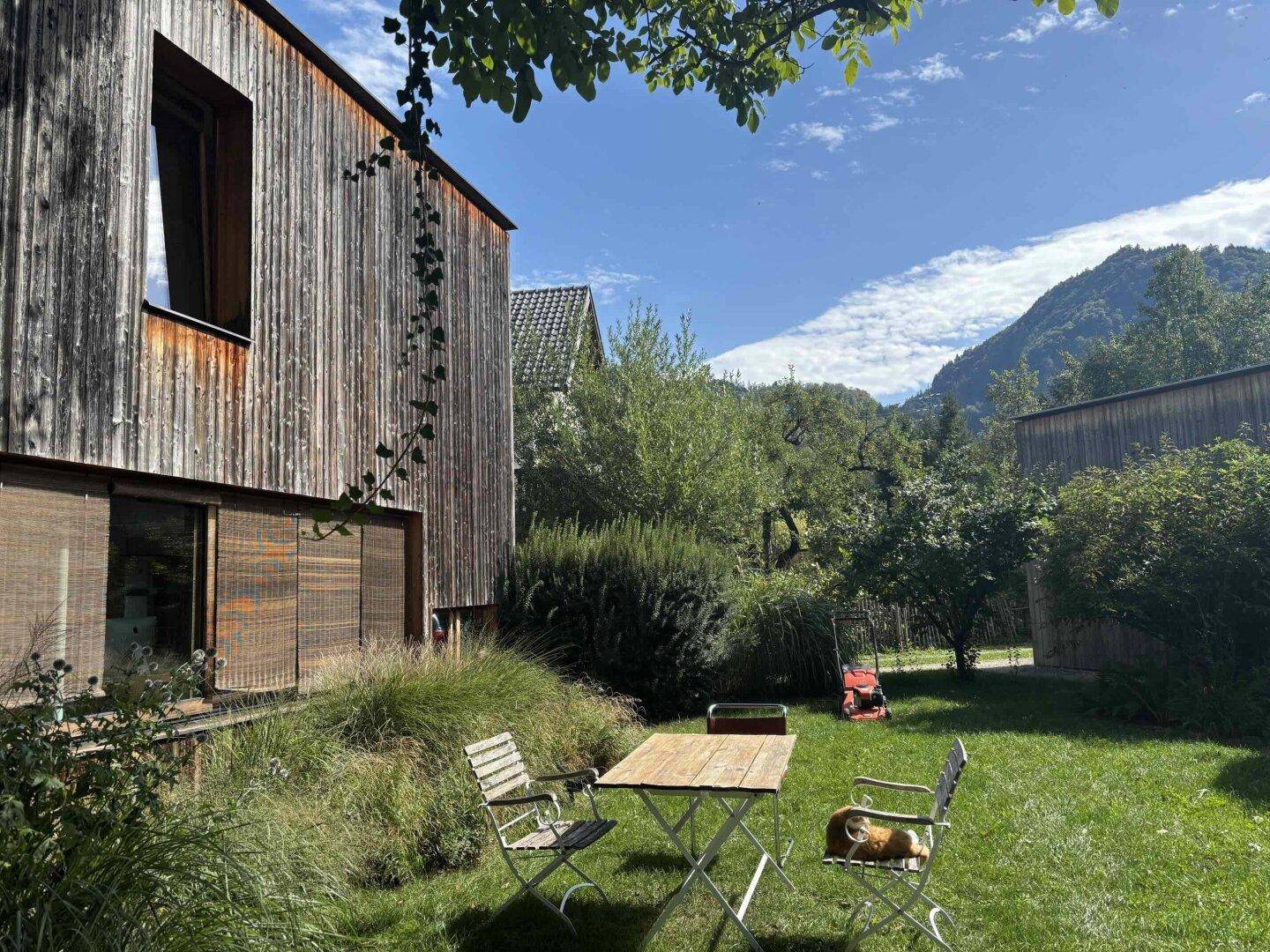 Gartenbereich mit Holztisch und Stühlen vor dem modernen Holzhaus mit Bergblick.