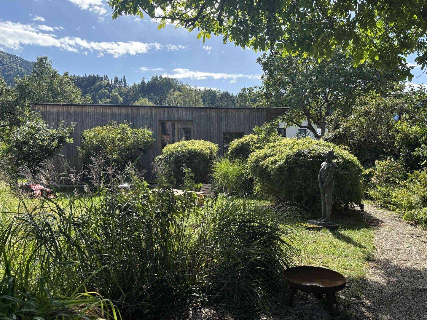 Weitläufiger Garten mit Skulptur und vielfältiger Bepflanzung vor dem modernen Holzhaus.