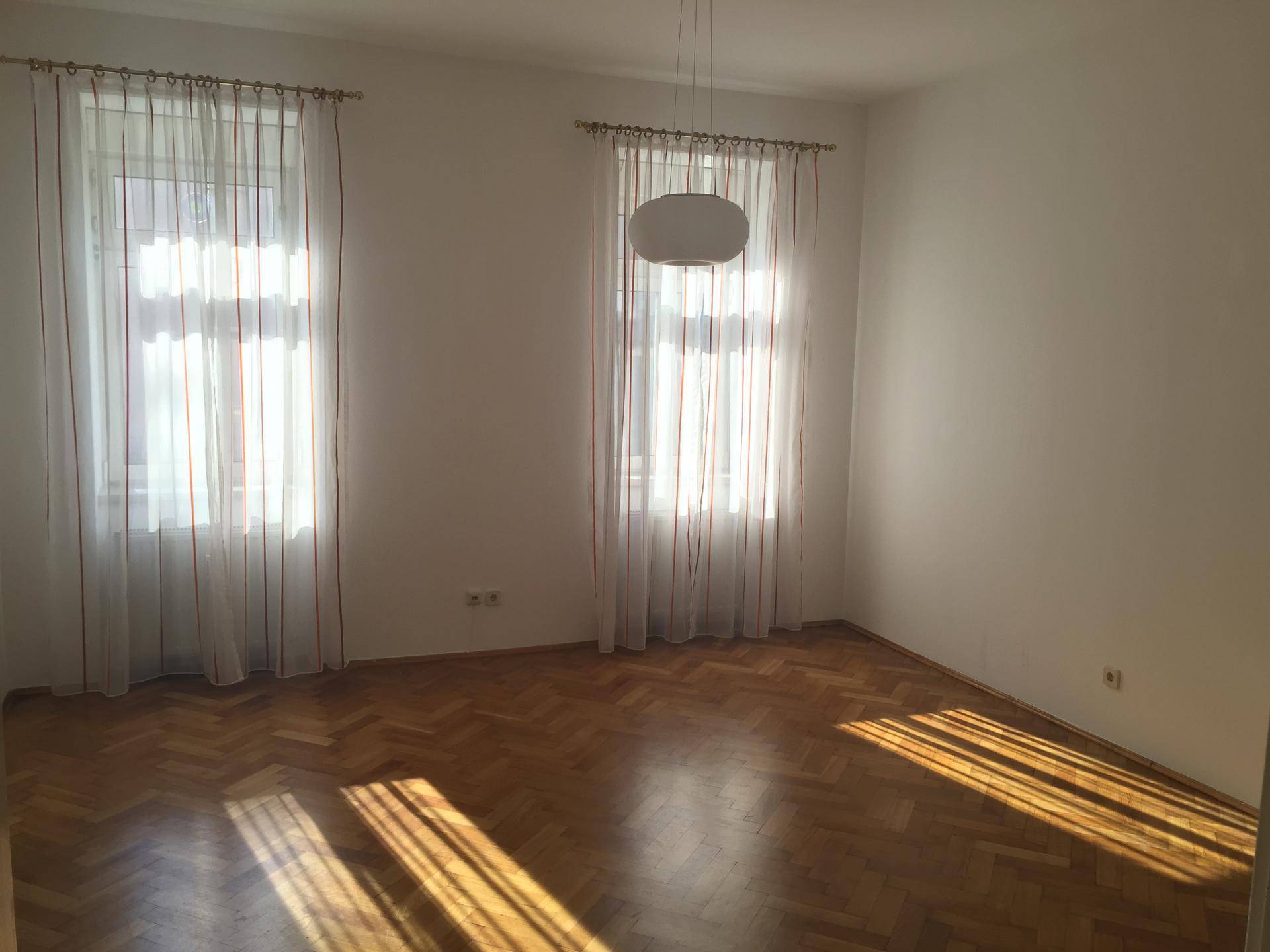 Geräumiges Zimmer mit zwei Fenstern, hellem Fischgrätparkett und viel Tageslicht.