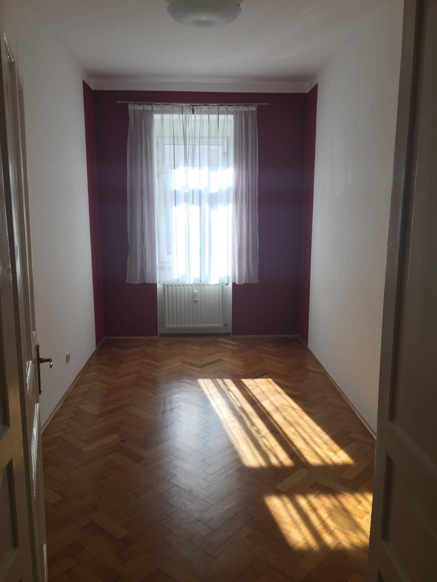Schmaler Raum mit roter Akzentwand, einem Fenster und klassischem Fischgrätparkettboden.