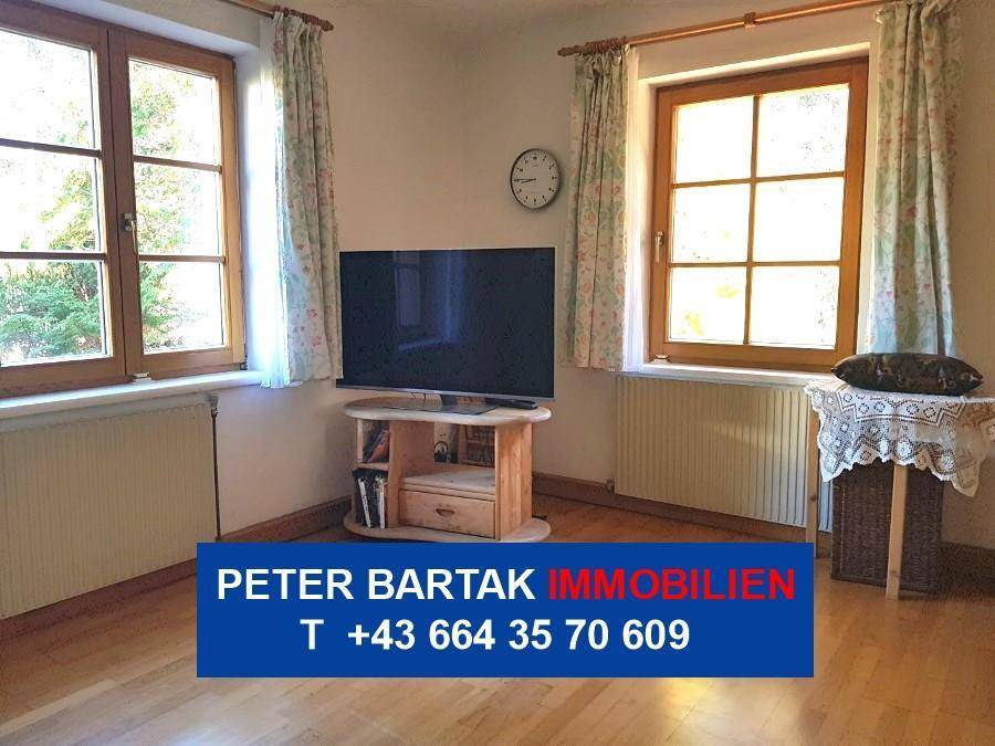 Gemütliches Wohnzimmer mit Fernseher, Holzmöbeln und zwei Fenstern.