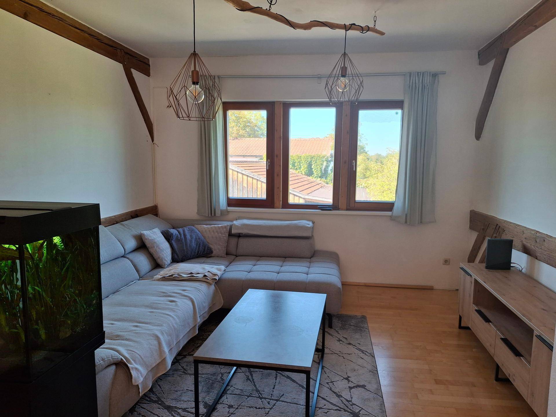Gemütliches Wohnzimmer mit großem Ecksofa, Couchtisch und Aquarium, beleuchtet durch ein Fenster mit Blick ins Grüne.