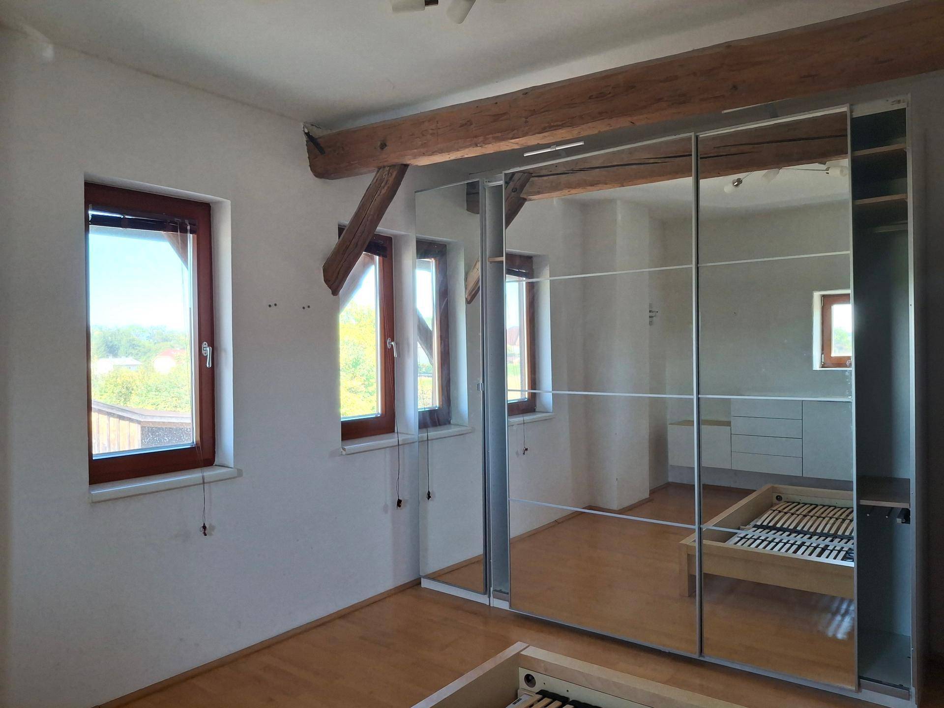 Schlafzimmer mit großem Spiegelschrank, Holzbalken und Fenstern, die viel Tageslicht hereinlassen.