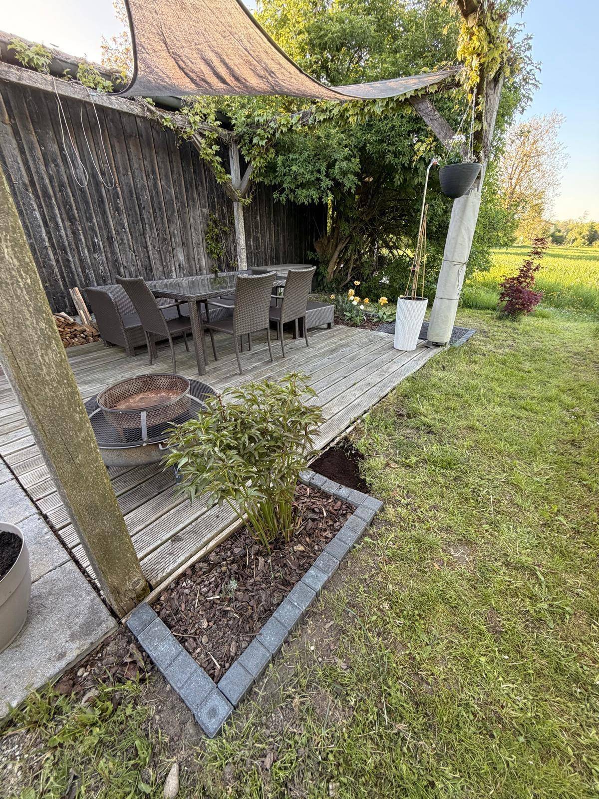 Gemütlicher Gartenbereich mit Holzterrasse, Sitzgelegenheiten und Feuerstelle, umgeben von viel Grün.