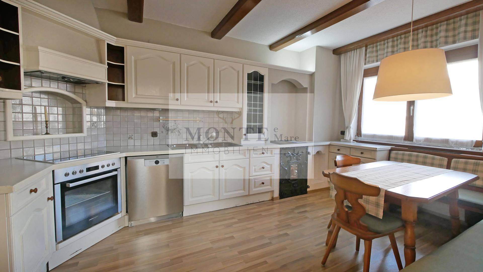 Helle Küche mit weißen Schränken, Holzbalken und Essbereich am Fenster.