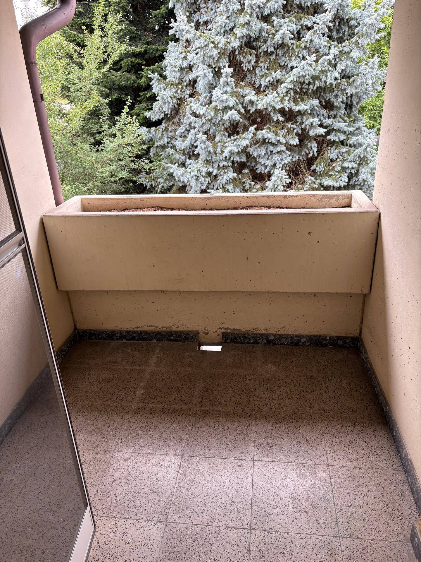 Kleiner Balkon mit Fliesenboden und integriertem Blumenkasten, umgeben von grünen Bäumen.