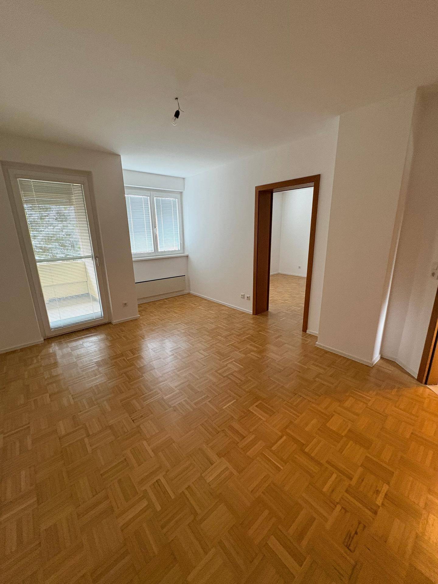 Geräumiges Wohnzimmer mit hellem Parkettboden, weißen Wänden und Zugang zum Balkon.