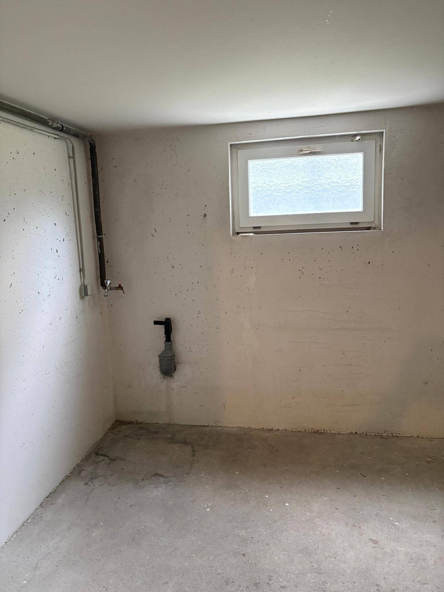 Heller Kellerraum mit kleinem Fenster, Betonboden und sichtbaren Installationen an der Wand.