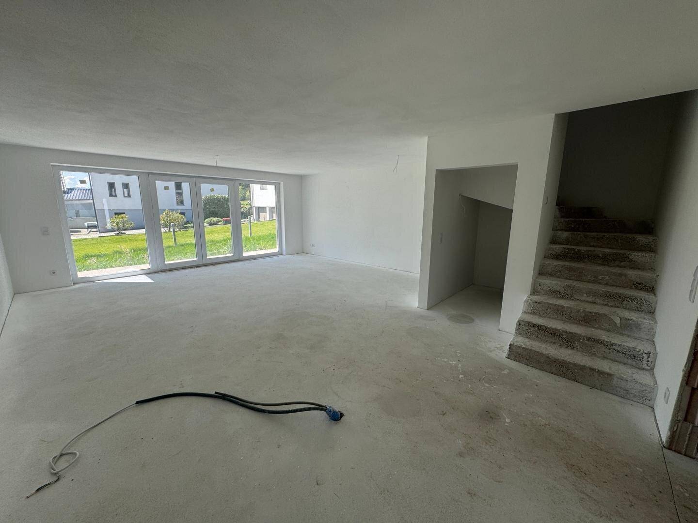 Geräumiger Wohnbereich im Rohbau mit großen Fensterfronten und Treppenzugang.