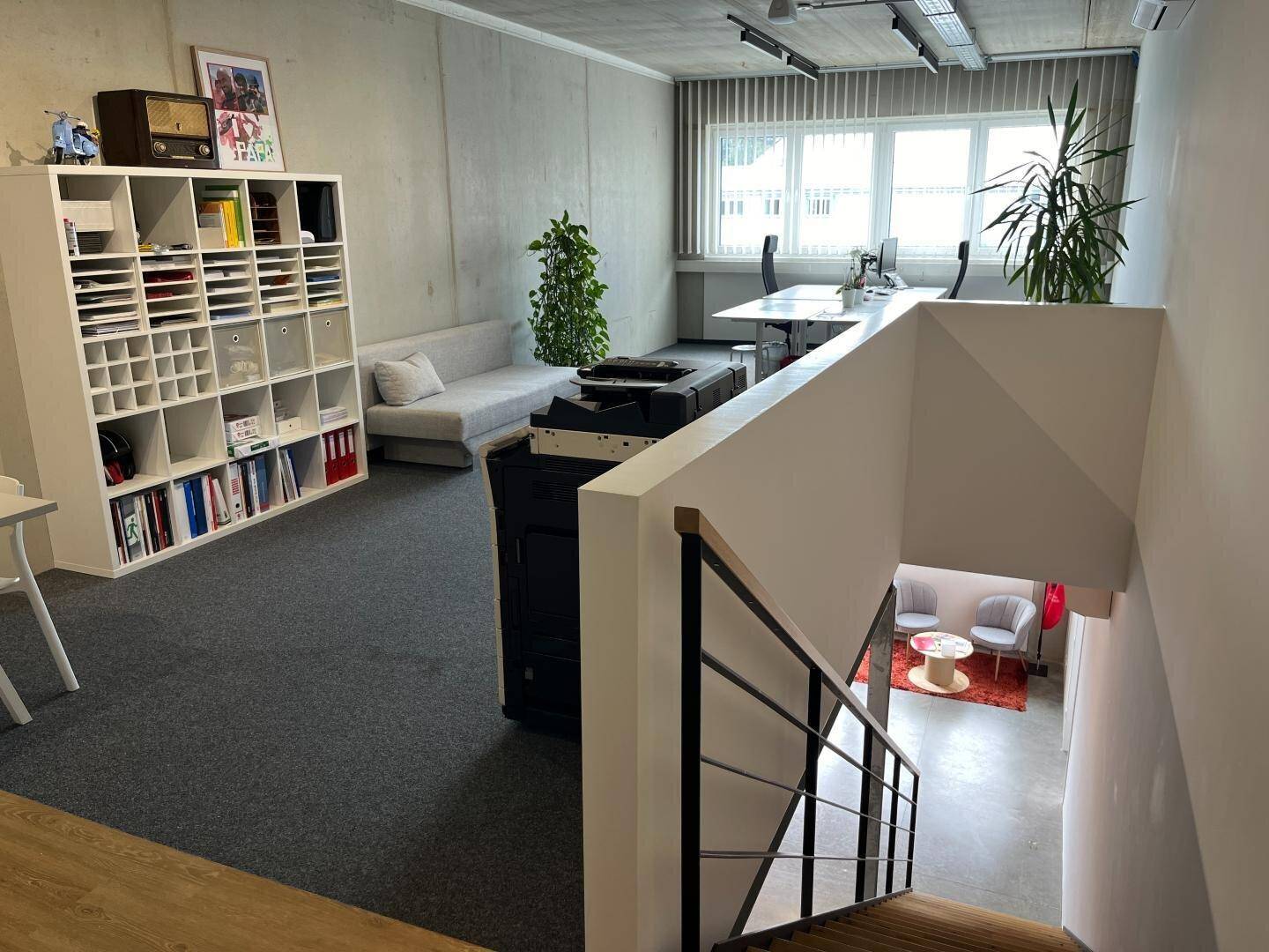 Helles Büro mit Teppichboden, Schreibtischen und einem Blick auf einen tiefer gelegenen Bereich mit Sitzgelegenheiten.