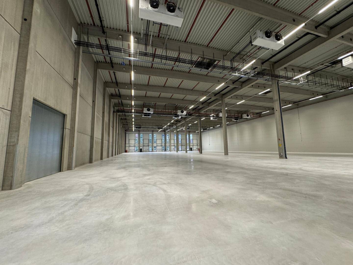 Weitläufige, leere Lagerhalle mit glattem Betonboden, hoher Decke und installierten Lüftungsanlagen.