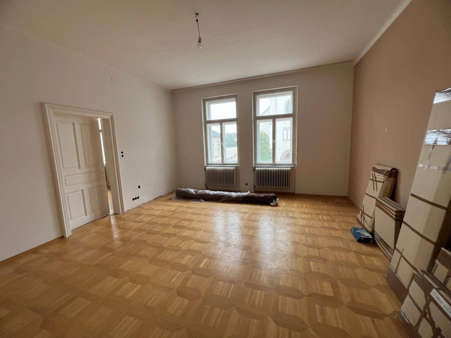 Geräumiges, leeres Zimmer mit Fischgrätparkett, hohen Decken und großen Fenstern, bereit für die Renovierung.