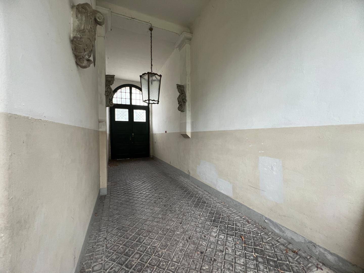 Langer, schmaler Eingangsbereich mit historischen Fliesen und einer hohen Decke, die zu einer dunklen Tür führt.