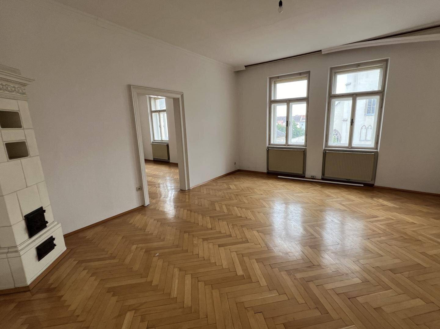 Geräumiges, leeres Zimmer mit Fischgrätparkett, einem traditionellen Kachelofen und großen Fenstern.
