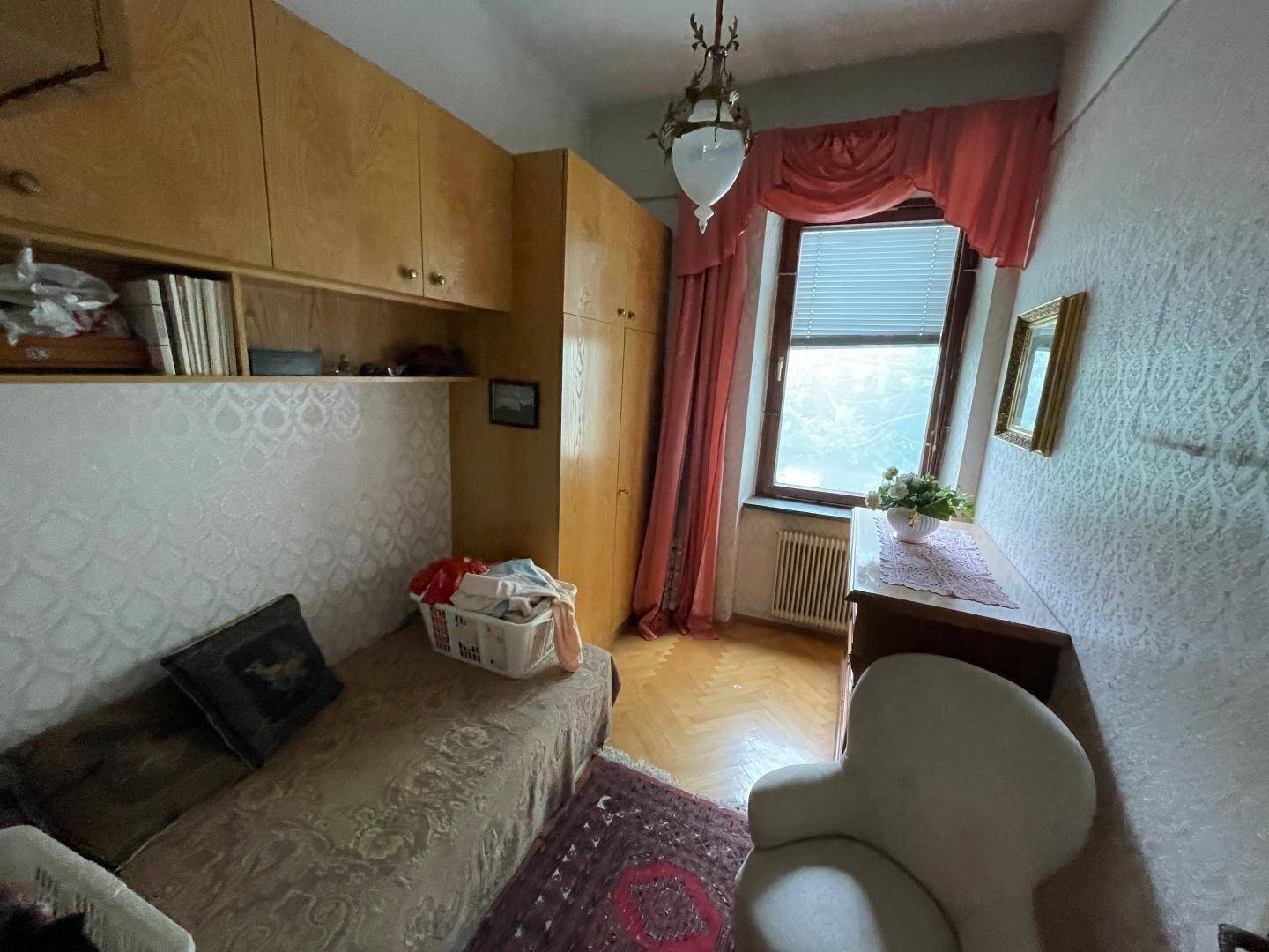Kleines Schlafzimmer mit Holzschränken, gemusterter Tapete und einem Fenster, das Tageslicht hereinlässt.