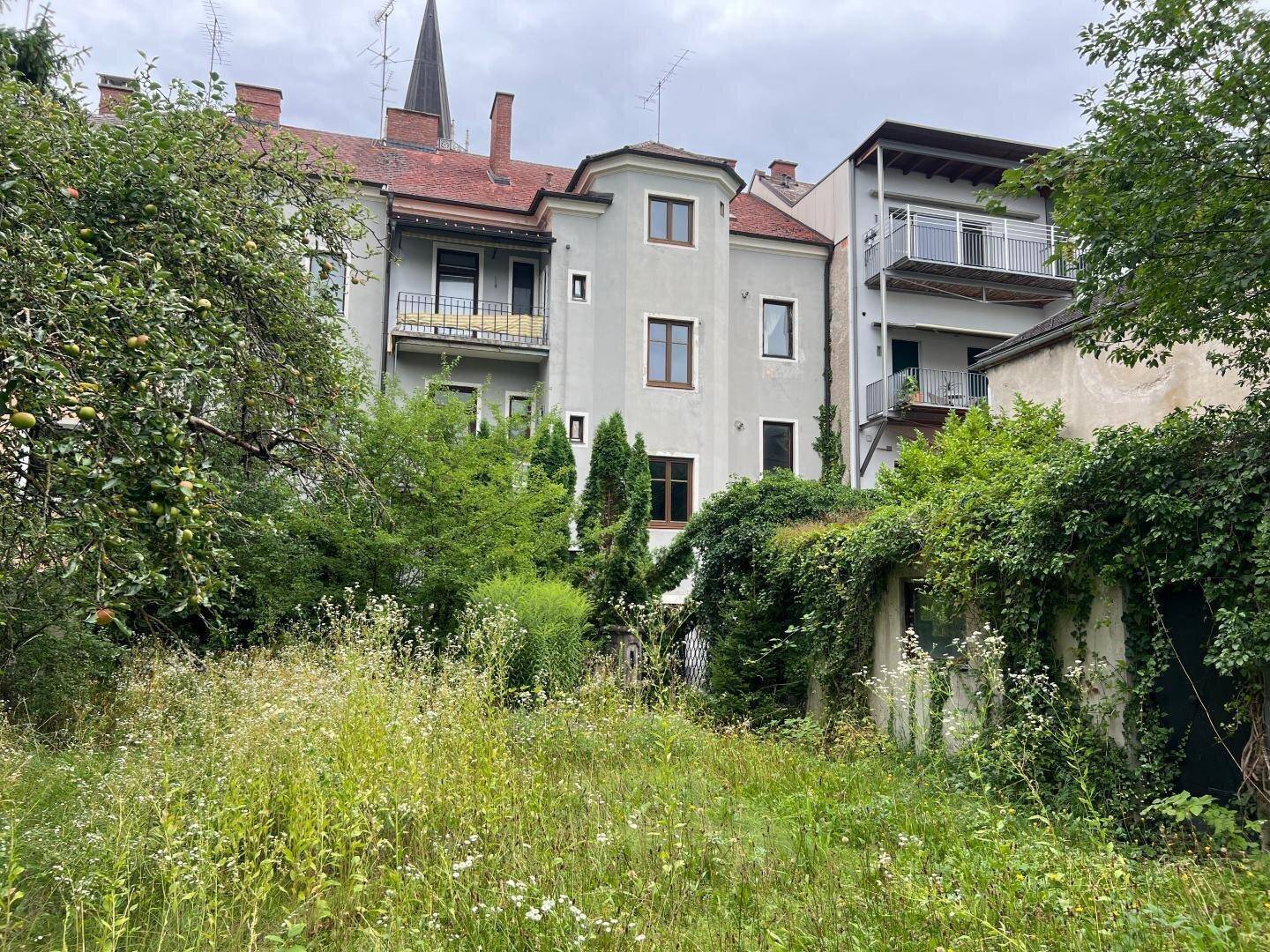 Hinteransicht eines Gebäudes mit Balkonen und einem großen, verwilderten Gartenbereich mit Bäumen.
