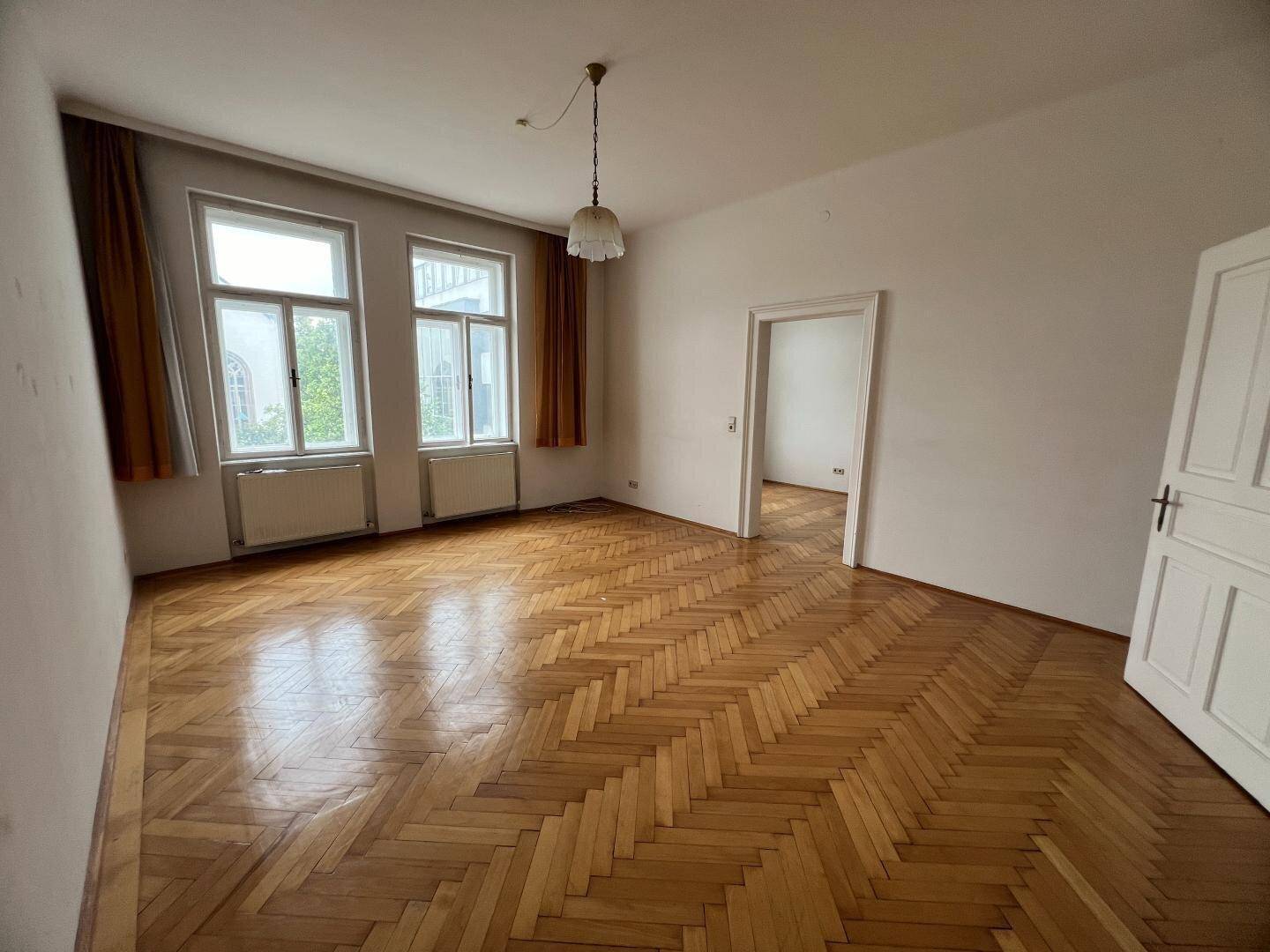 Helles, leeres Zimmer mit Fischgrätparkett, hohen Decken und großen Fenstern, die viel Licht hereinlassen.