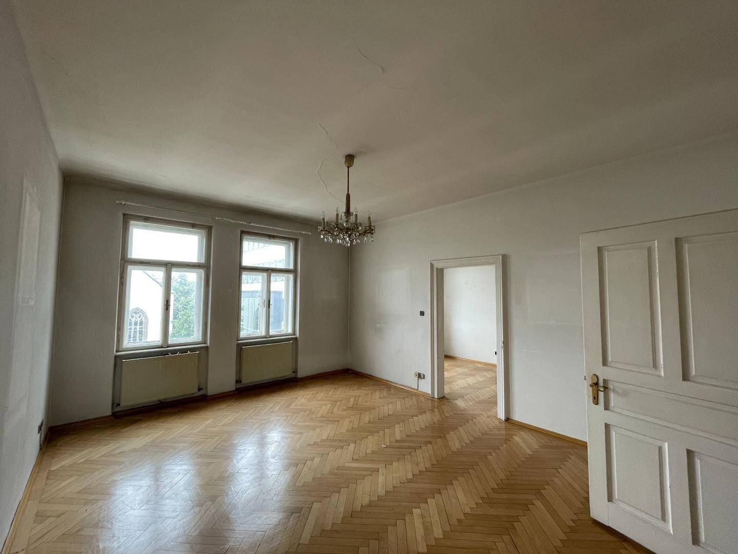 Helles, leeres Zimmer mit Fischgrätparkett, hohen Decken und großen Fenstern, die viel Licht hereinlassen.