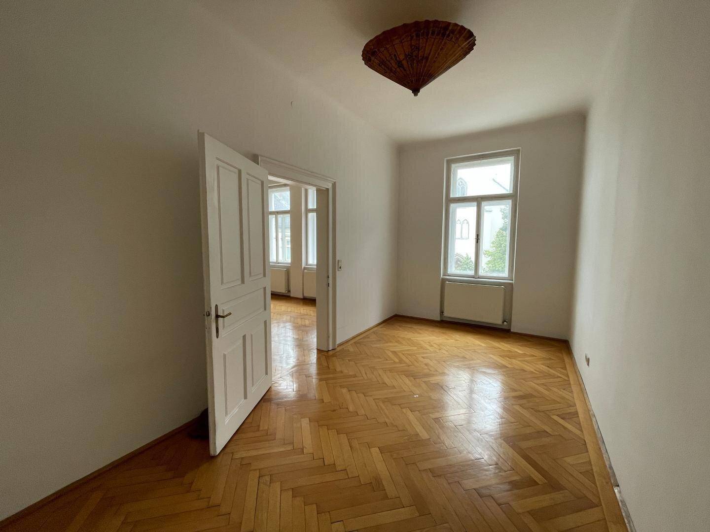 Leeres Zimmer mit Fischgrätparkett, hohen Decken und einem großen Fenster, das den Raum erhellt.