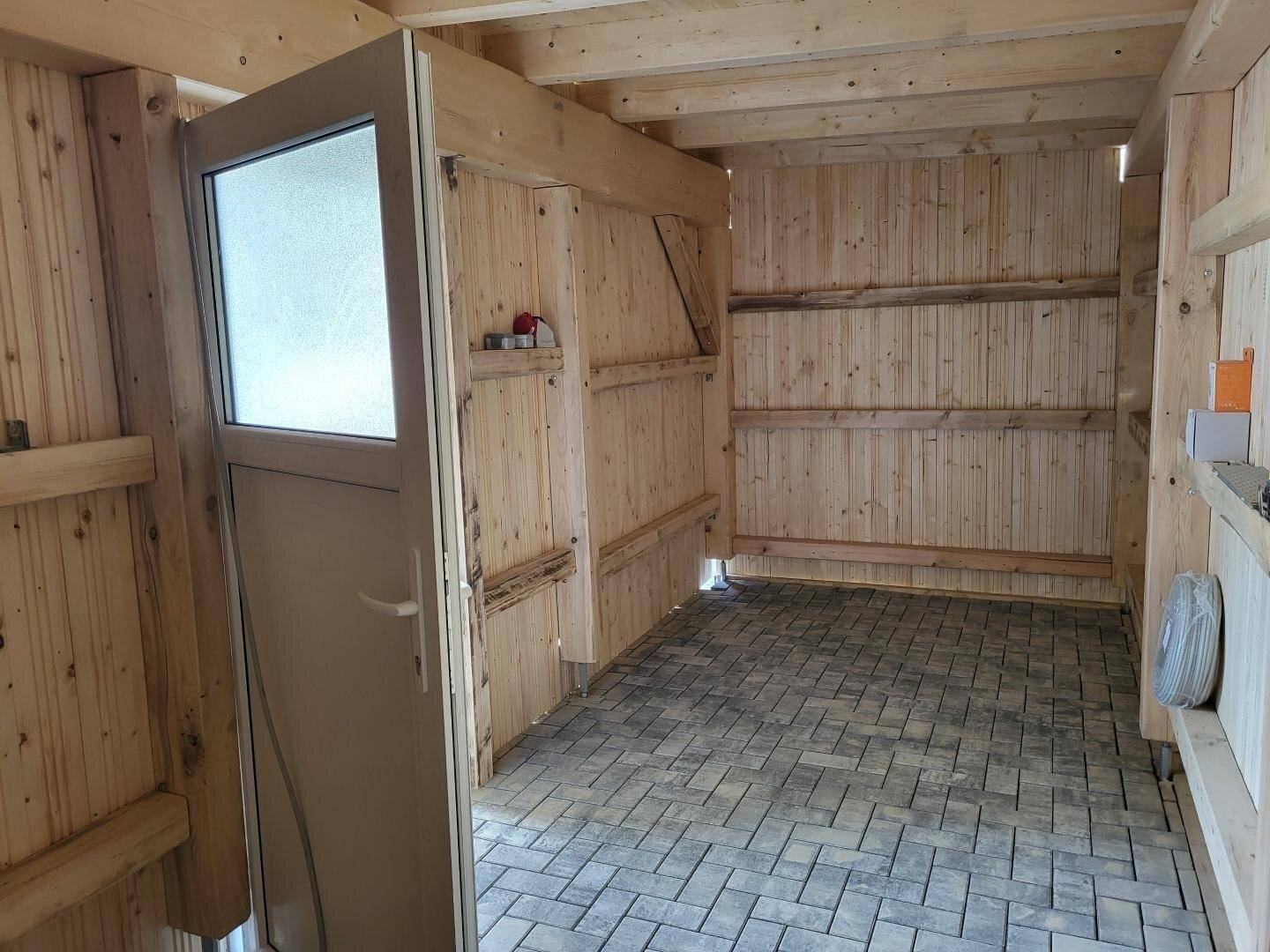 Abgeschlossener Abstellraum im Carport mit Holzwänden und gepflastertem Boden.