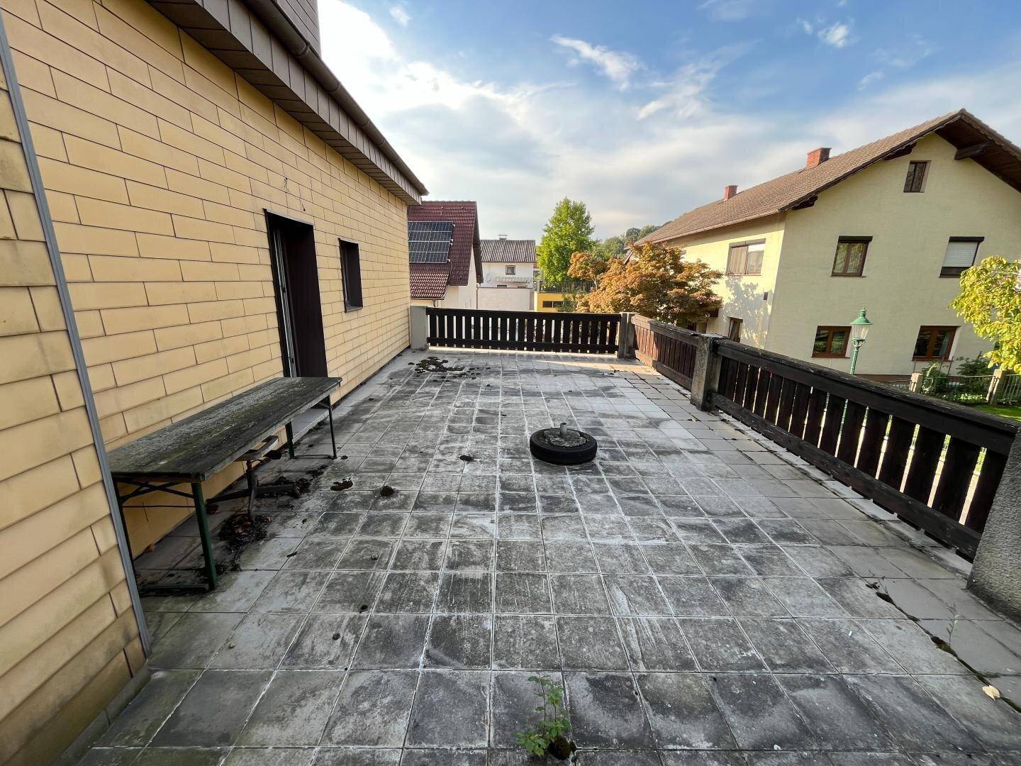 Weitläufige Außenterrasse mit Fliesenboden und Holzgeländer, umgeben von Grün.