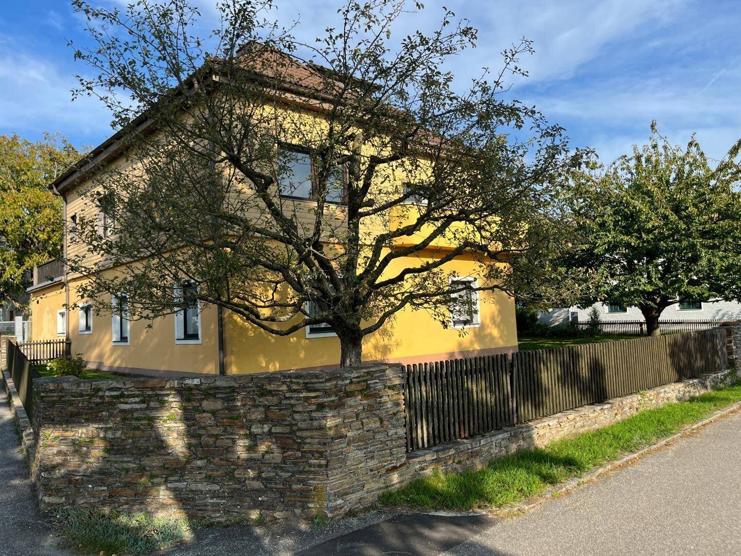 Seitenansicht des gelben Hauses mit Steinfundament und Holzzaun unter blauem Himmel.