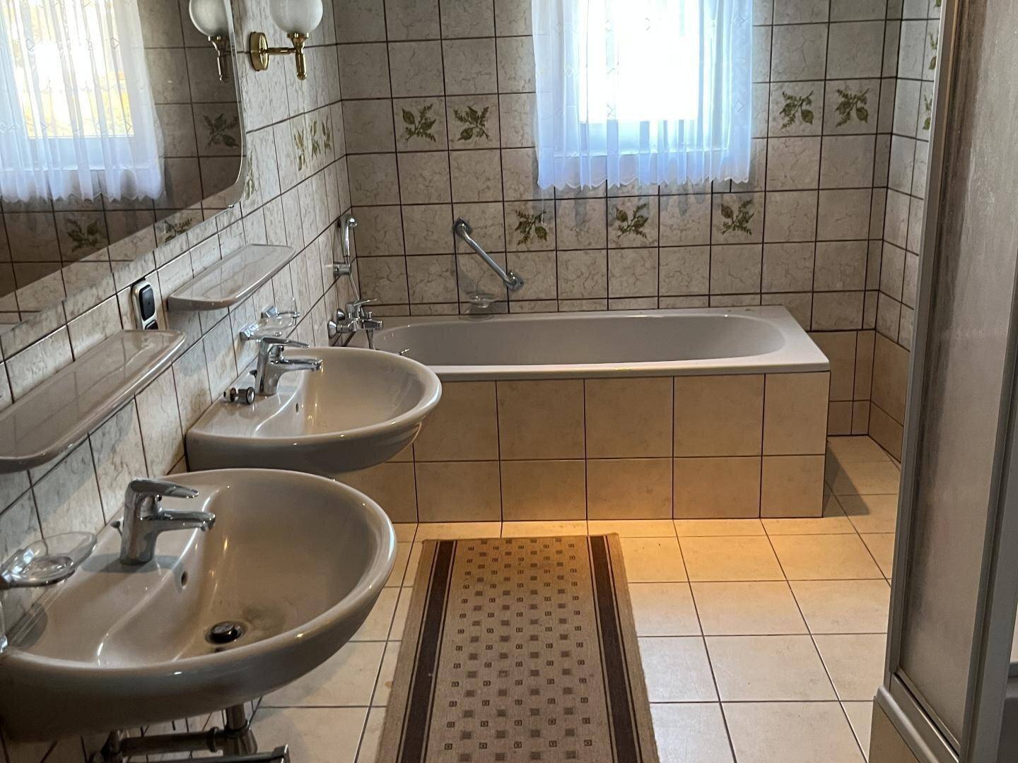 Badezimmer im 80er-Jahre-Stil mit zwei Waschbecken, Badewanne und gemusterten Fliesen.