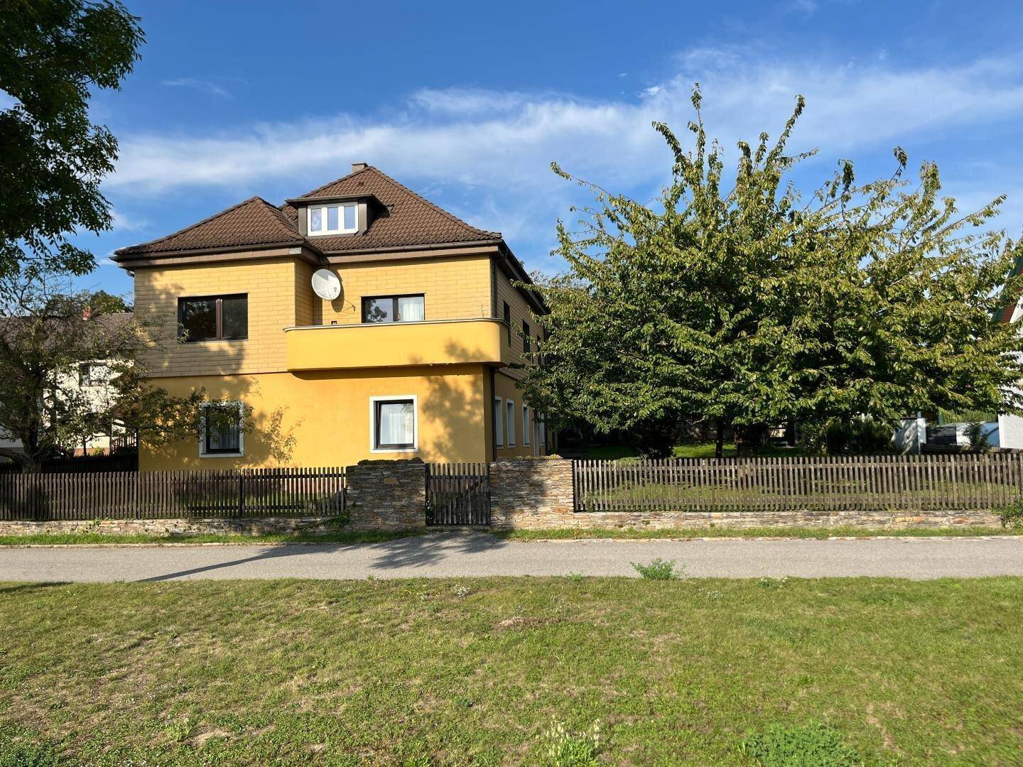 Ansicht des gelben Hauses mit braunem Dach, umgeben von einem Holzzaun und Bäumen.
