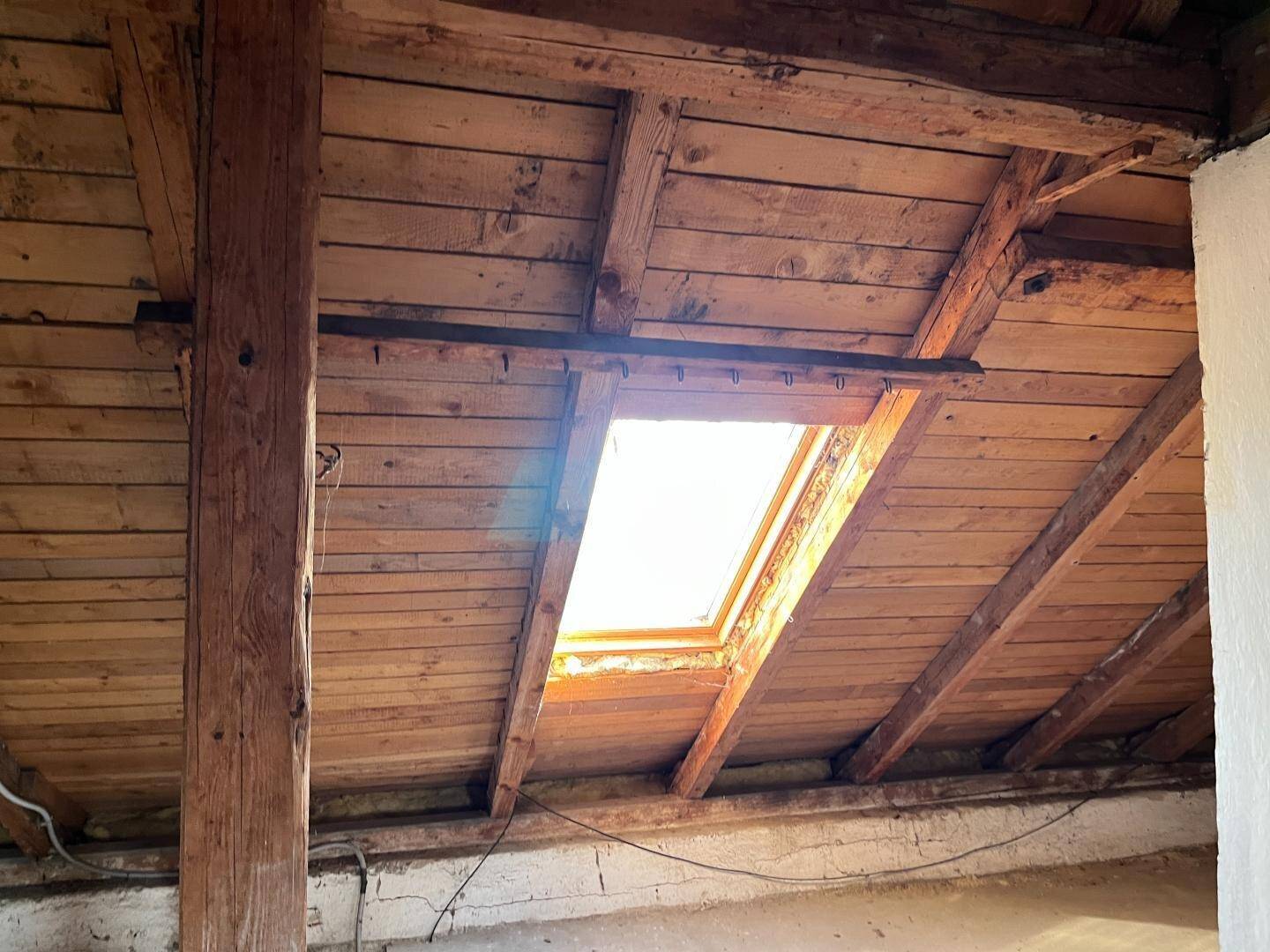 Dachboden mit sichtbaren Holzbalken und einem Dachfenster, das Tageslicht hereinlässt.