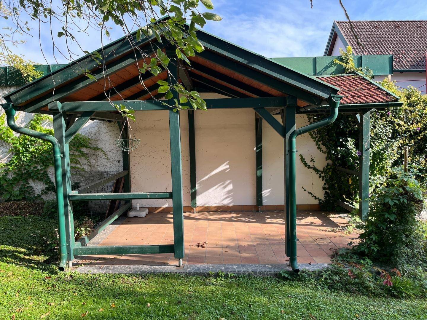 Überdachter Sitzbereich im Freien mit Fliesenboden und grünem Holzrahmen.